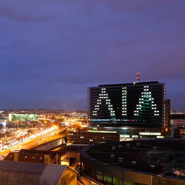 Photo de nuit d'un immeuble au milieu d'une ville éclairée, avec les lettres AIA en grand, formées par les fenêtres éclairées sur la façade de l'immeuble