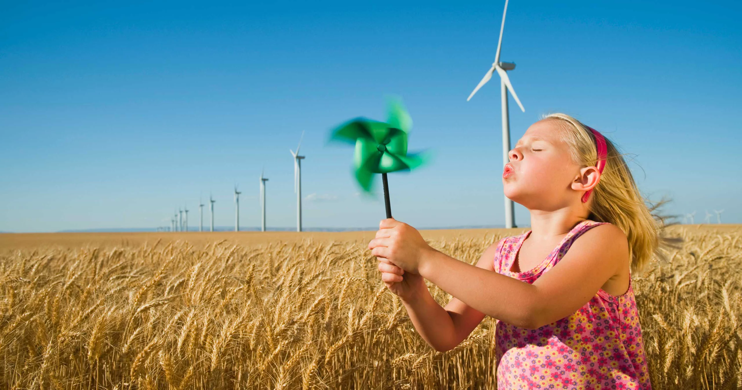 Photo d'une petite fille dans un champ de blé soufflant sur un jouet moulin à vent, avec des éoliennes en fond