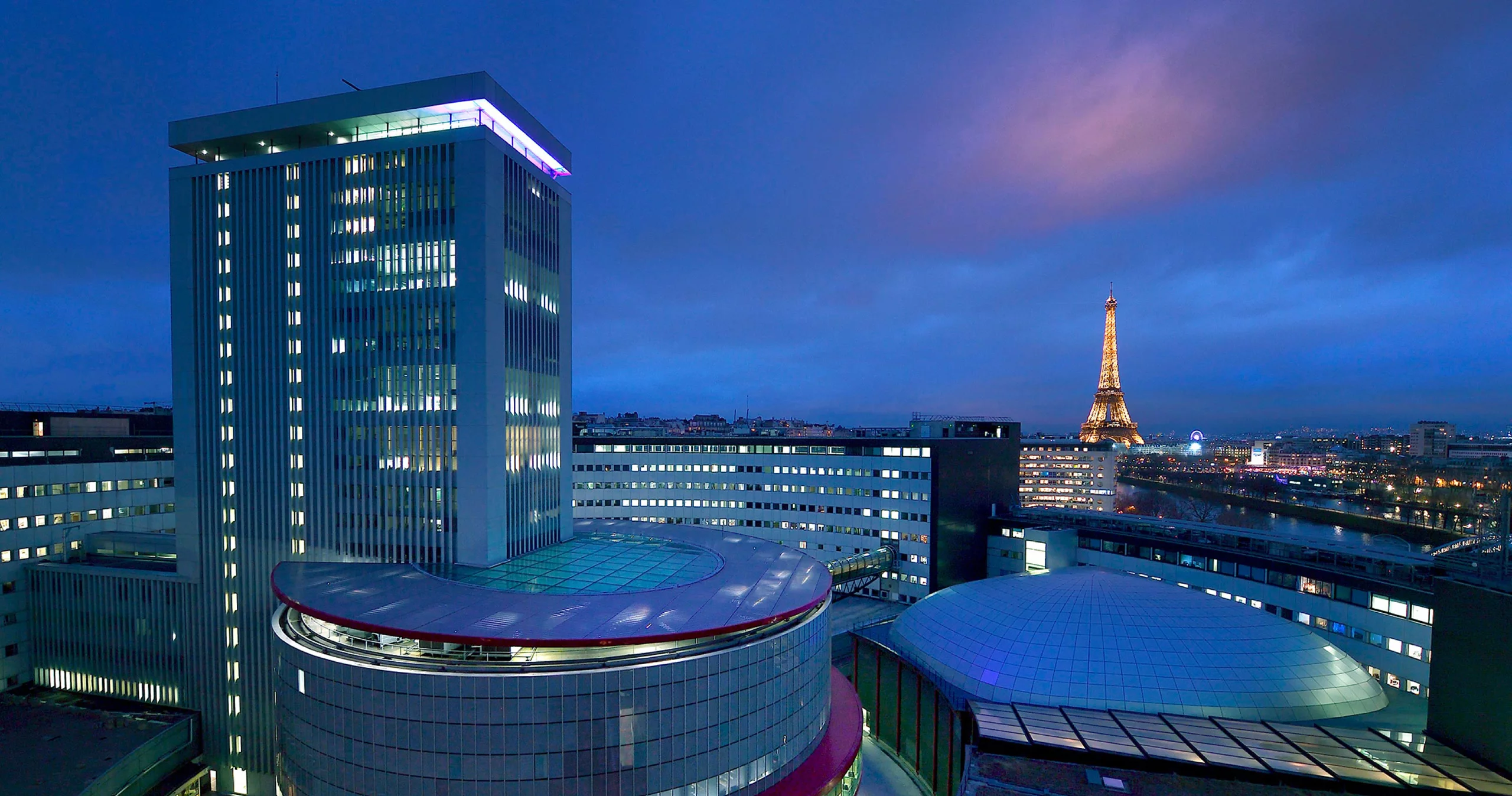 Photo des bâtiments modernes de Radio France éclairés le soir, avec la ville et la tour Eiffel éclairées en arrière-plan