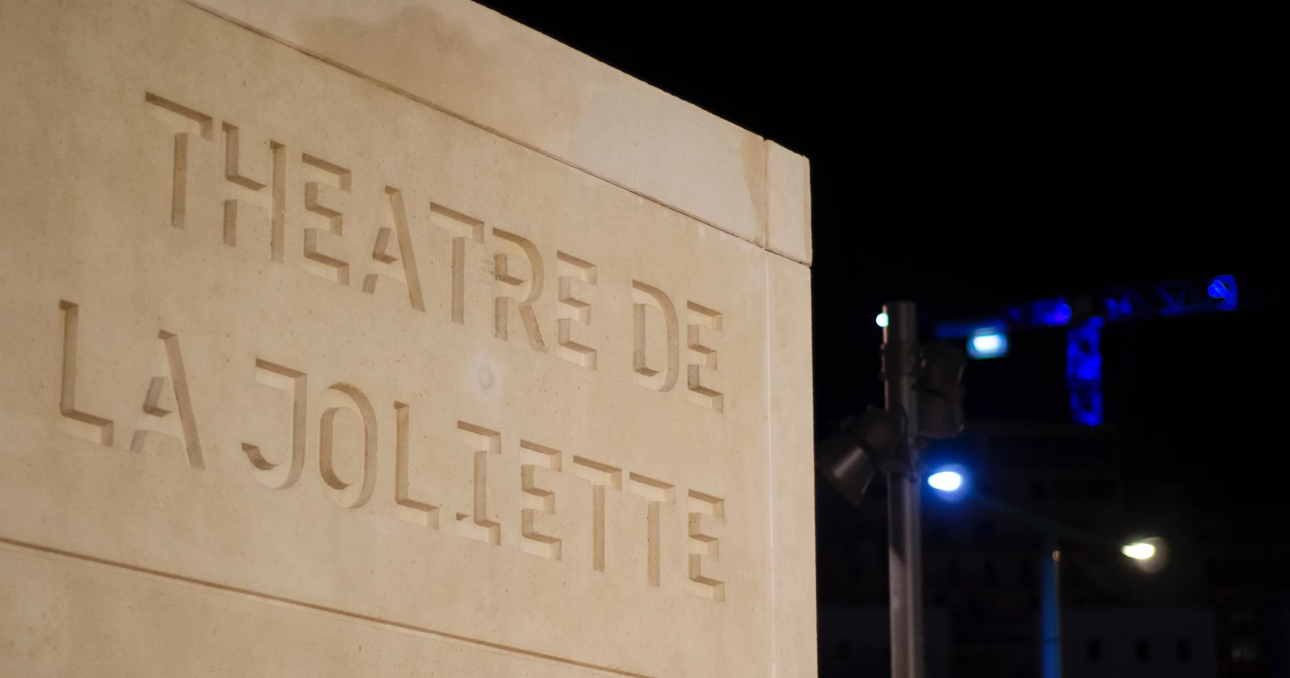 Photo du nom "Théâtre de la Joliette" gravé dans de la pierre avec une scène de spectacle dans l'obscurité en arrière-plan