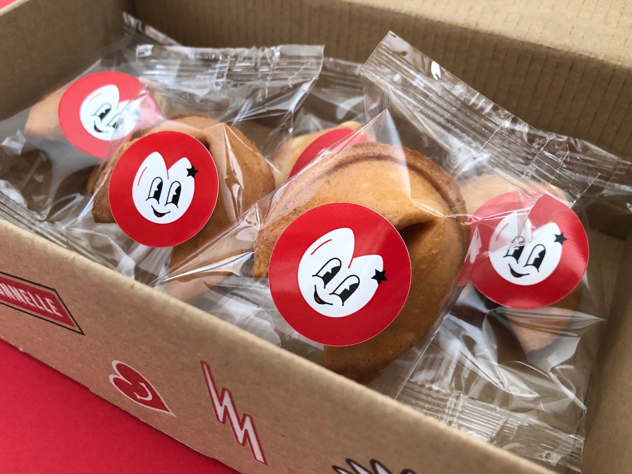des fortunes cookies dans des emballages individuels avec un sticker rouge, noir et blanc qui représente un personnage en forme de fortune cookie. Le tout dans la boite en kraft décorée.